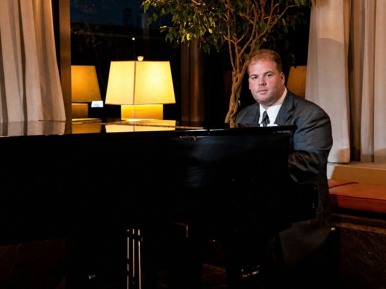 David Spatola in a suit at a grand piano in a warm, lamplit hotel lobby
